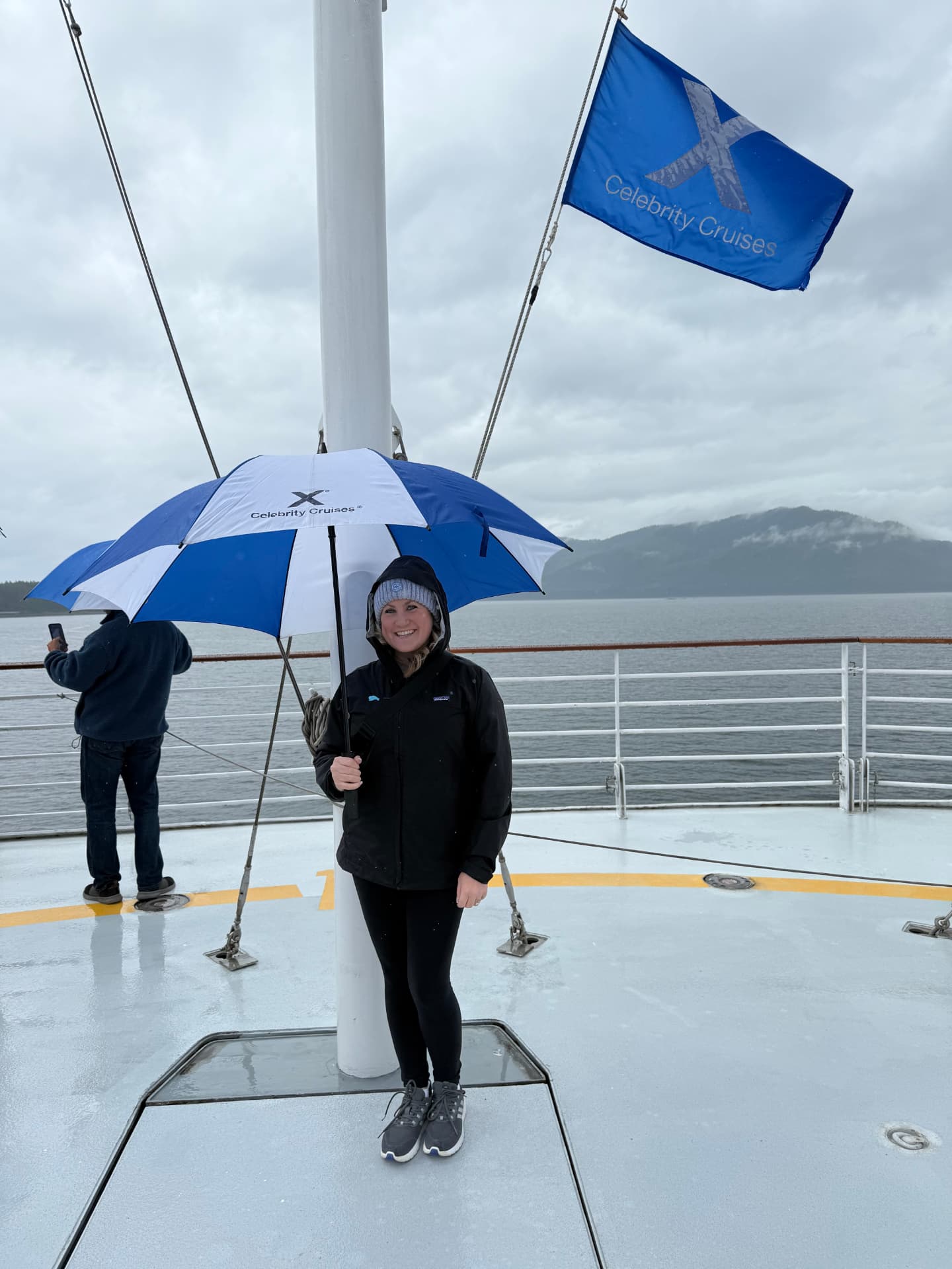 A guest under a Celebrity Cruises umbrella on deck with misty mountains behind