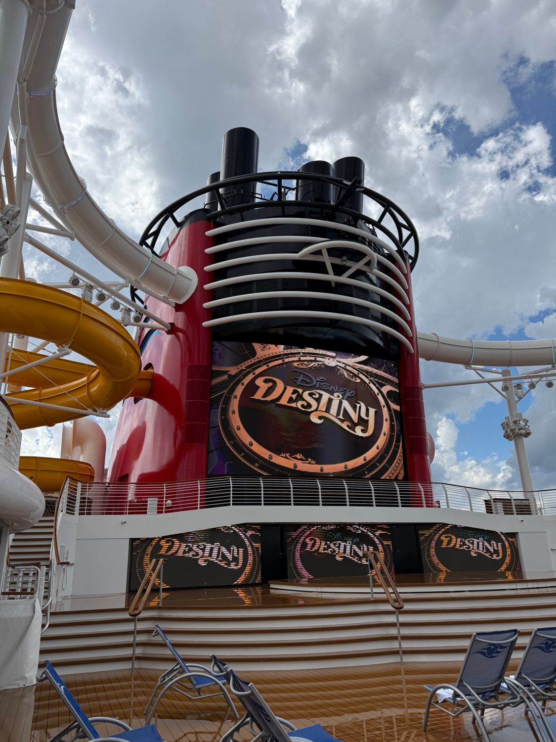 The funnel and pool deck of the Disney Destiny, with a tall red funnel fitted with screens, water slides and lounge chairs