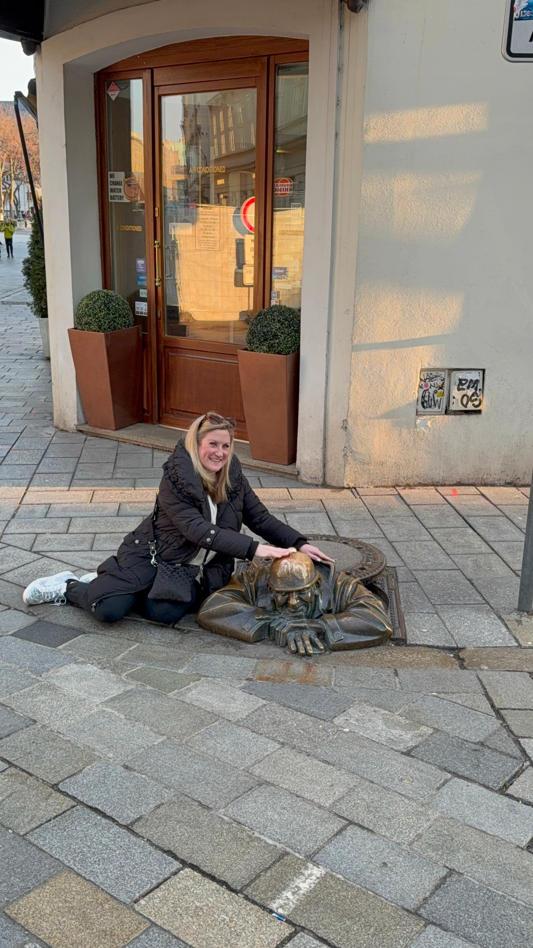 Travel advisor Bridget Edwards of Illumia Travel patting Čumil, the bronze statue of a man peeking out of a manhole in Bratislava's Old Town