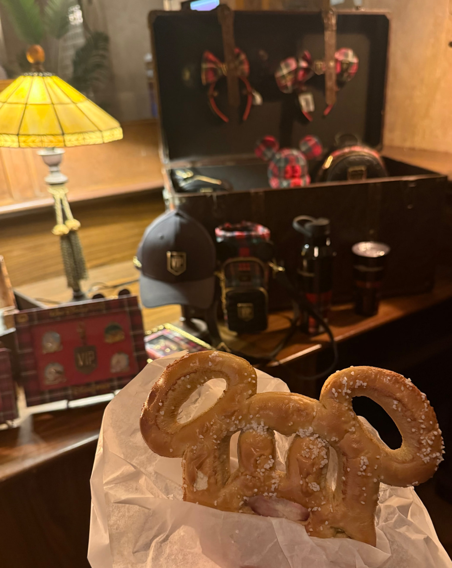 A Mickey-shaped pretzel held in front of a trunk of plaid Minnie ears, a VIP cap and VIP pins